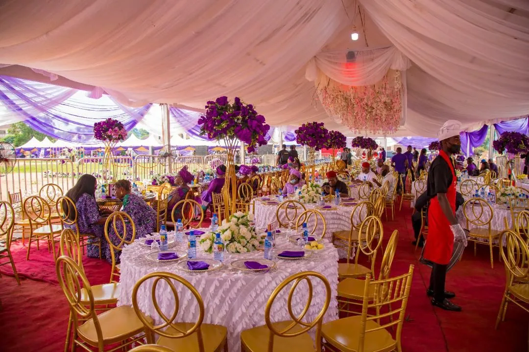 Formal event tent with white draping, purple flowers, and gold seating